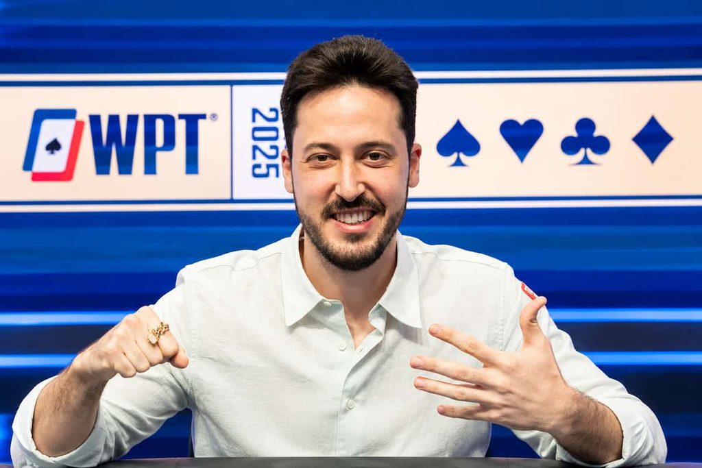 Adrian Mateos smiling at a poker table during a tournament, wearing a white shirt with a blue poker-themed backdrop behind him.