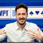 Adrian Mateos smiling at a poker table during a tournament, wearing a white shirt with a blue poker-themed backdrop behind him.